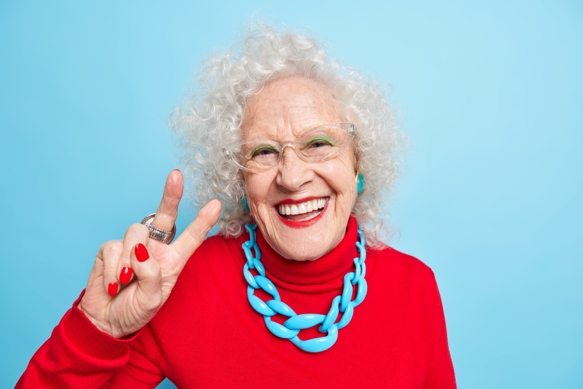 An elderly smiling and joyful woman showing the peace sign wearing a red sweater