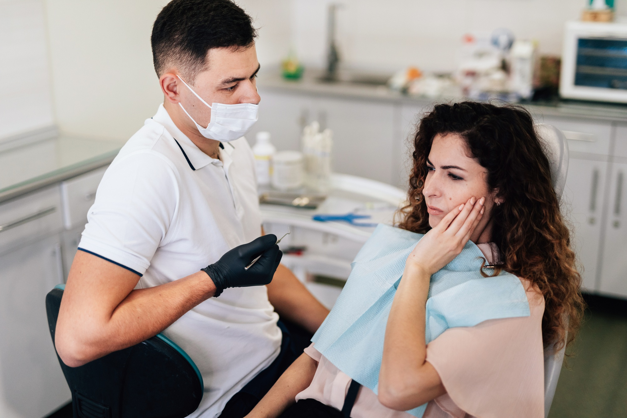 A young woman at the dentist's office and chair holding a hand to the side of her face and looking worried.